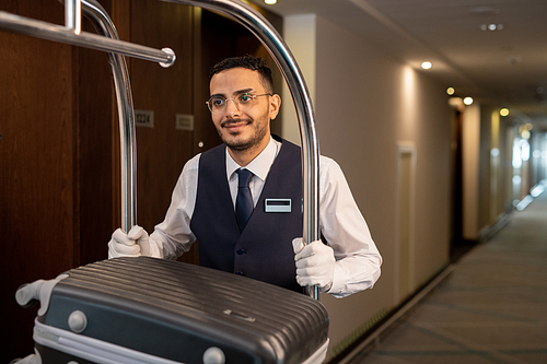 Young smiling porter in uniform and eyeglasses pushing cart with suitcases of guests while moving in front of camera along corridor in hotel