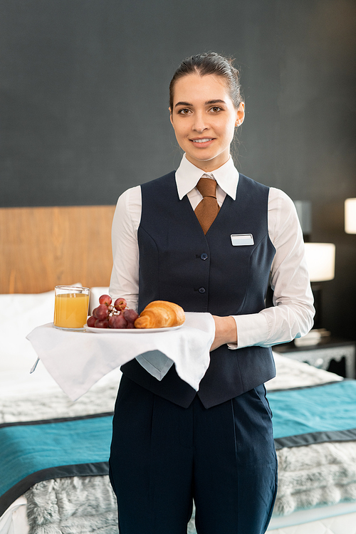 Happy young elegant female worker of hotel holding tray with breakfast while standing in front of camera against bed and looking at you
