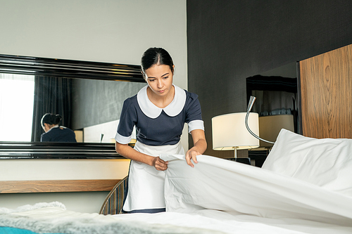 Young pretty brunette maid putting white clean blanket while changing bed in one of hotel rooms during preparation for new guests