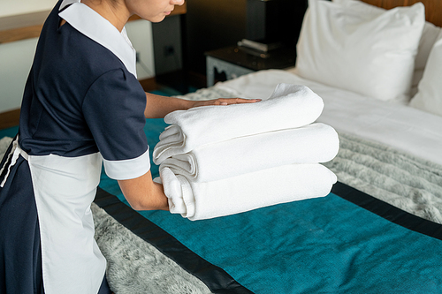 Young beautiful chambermaid in uniform holding stack of clean folded towels while going to put them on changed bed in hotel room