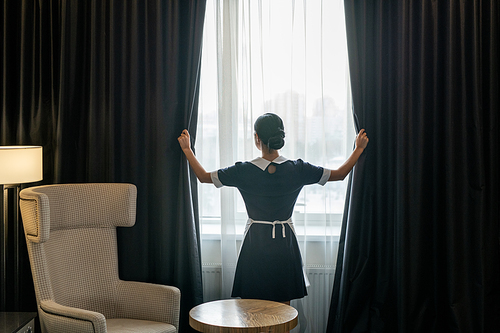 Back view of young chambermaid in uniform opening dark curtains hanging on large window of hotel room before cleaning it