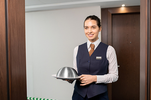 Happy young elegant manager in uniform holding tray with breakfast while standing in front of open door in the corridor of luxurious hotel
