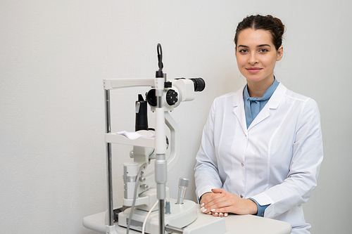 Young smiling female ophthalmologist in whitecoat sitting in front of camera by medical equipment in clinics and waiting for patient
