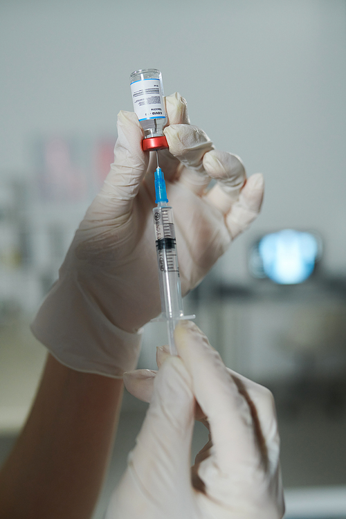 Gloved hands of clinician or nurse holding syringe and small bottle with new vaccine while preparing injection for patient with covid19