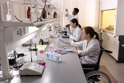 Young woman sitting in wheelchair and working with test tubes with her colleagues working in the background