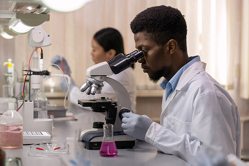 African young scientist sitting at the table looking through the microscope and examining samples in the laboratory