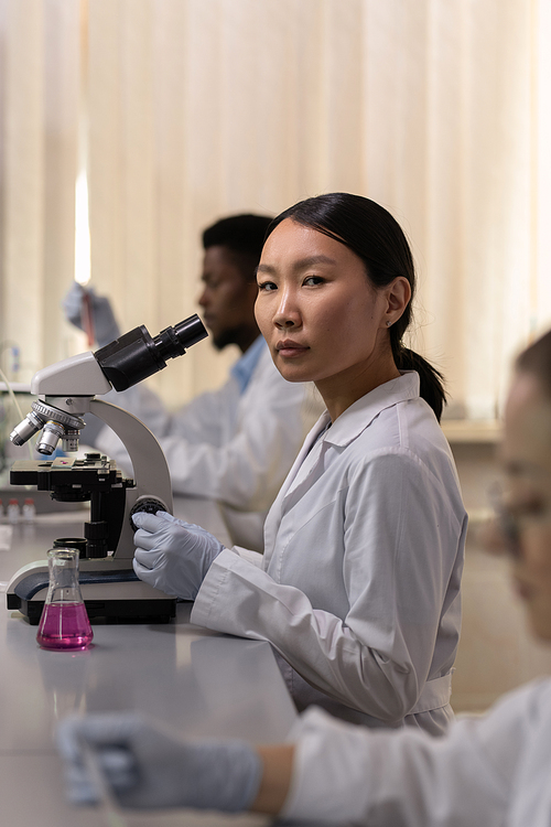 Portrait of Asian chemist sitting at the table and examining samples using microscope in the lab