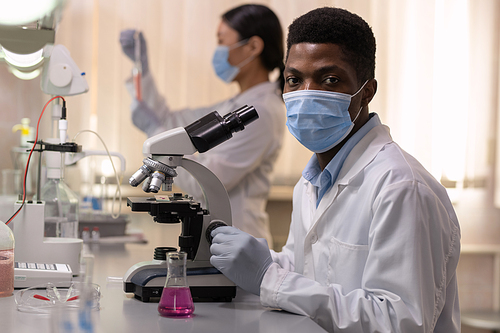 Portrait of African young chemist in mask looking at camera while working with microscope with his colleagues in the lab