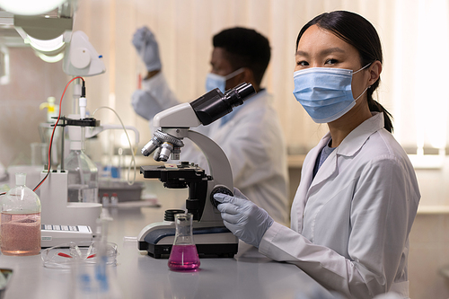 Portrait of Asian young scientist in protective mask looking at camera while working with microscope at the table in the lab