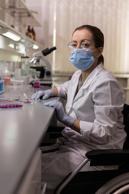 Portrait of Asian young chemist in mask sitting in wheelchair and looking at camera while working with samples in test tubes