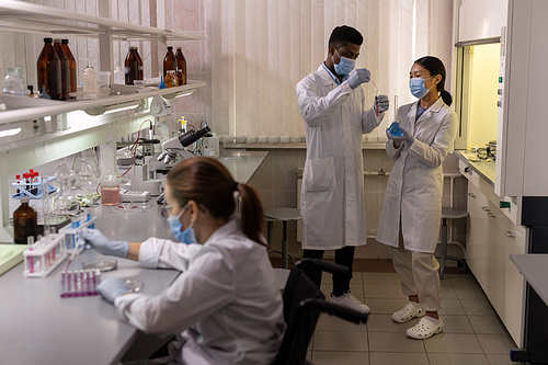 Group of chemists in white coats and masks examining samples and test tubes during teamwork in the laboratory