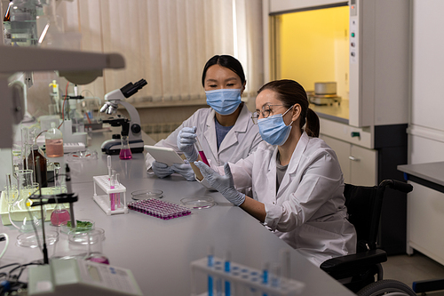 Young woman in mask examining liquid in test tube with her colleague sitting near by and using digital tablet they working together in the lab
