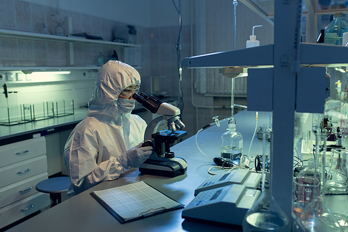 Scientist in protective workwear examining bacterium through the microscope at the table in the lab