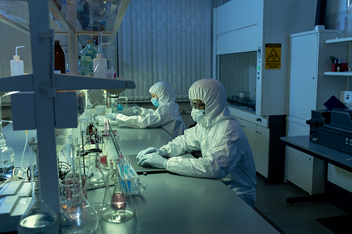 Two chemists in protective workwear sitting at the table and working on computers in the laboratory