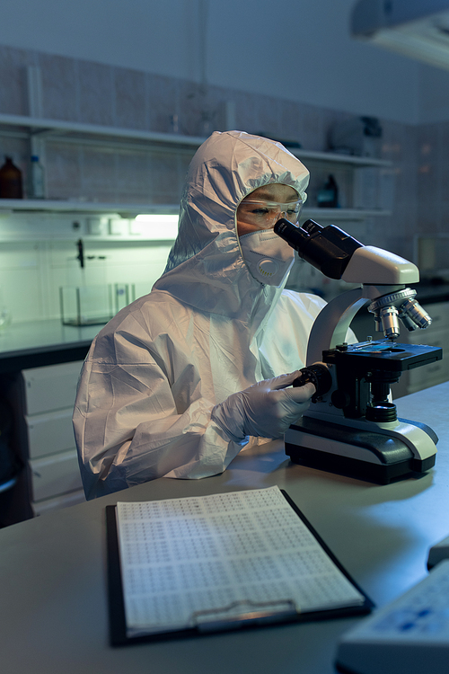Female chemist in protective wear sitting at her workplace and looking through the microscope while working in the lab