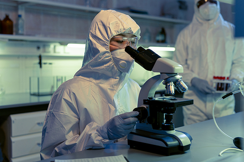 Scientist in protective workwear examining samples through the microscope at the table in the chemical laboratory