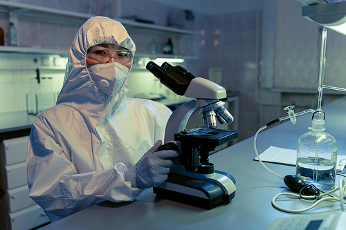 Portrait of female scientist in protective workwear looking at camera while working with microscope in chemical laboratory