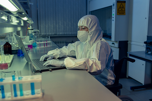 Female scientist in protective workwear sitting at the table and typing on laptop  during her work in the lab