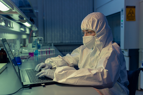 Scientist in protective workwear and mask typing on laptop at her workplace she working till late night in the lab