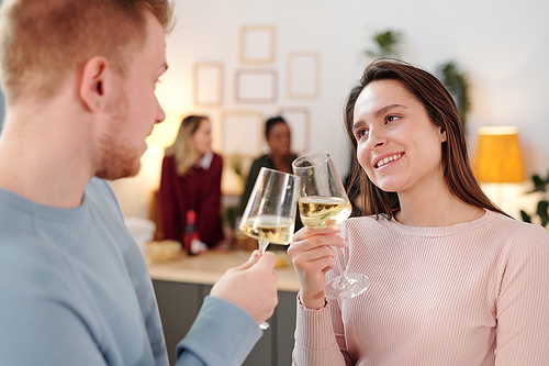 Happy young brunette woman in pink pullover clinking flute of champagne with that of her boyfriend while enjoying home party with friends