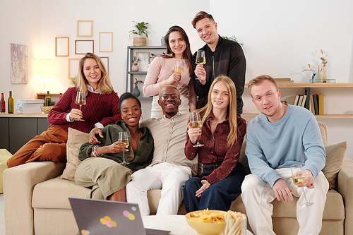 Group of young cheerful intercultural friends in smart casualwear sitting on large couch in living-room in front of camera and toasting