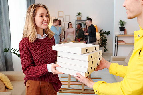 Young smiling blond female in smart casualwear taking stack of packed square carton boxes with pizza from hands of male courier in uniform