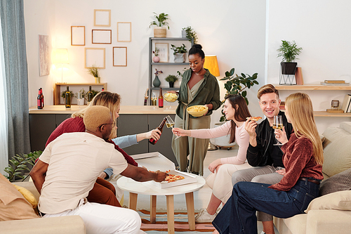 Group of young cheerful intercultural friends in smart casualwear sitting by small white round table in living-room, having pizza and drinks