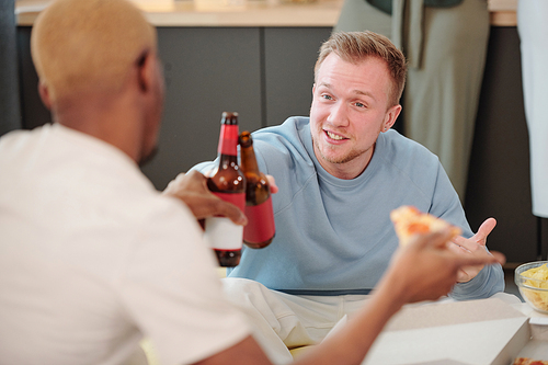 Happy blond guy in light blue sweatshirt looking at young African man while both clinking with bottles of beer over table and having snacks