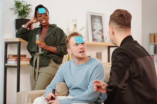 Two guys in casualwear looking at one another while trying to guess their names written on blue sticky notepapers during leisure game at party