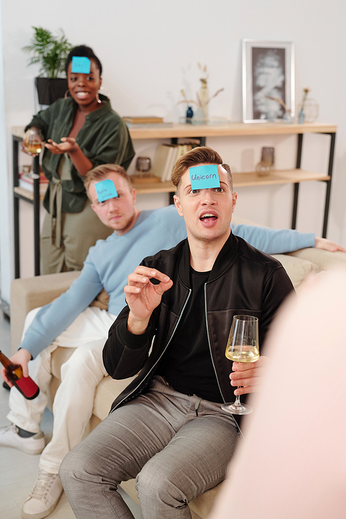 Young man with flute of champagne looking at one of friends while trying to guess his name written on blue sticky notepaper during leisure game