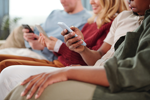 Group of young contemporary intercultural friends sitting in row on couch and scrolling in their mobile gadgets, focus on African couple