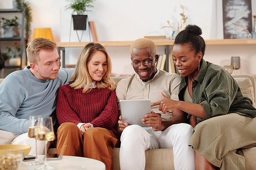 Group of young cheerful intercultural friends sitting in row on couch and discussing photos in social networks on screen of mobile gadget