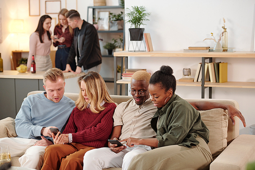 Two young intercultural couples in smart casualwear sitting in row on couch and discussing photos in social networks in mobile gadgets