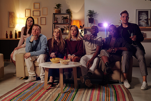 Group of young intercultural friends sitting on long couch by small table with snacks and watching movie together in living-room at party