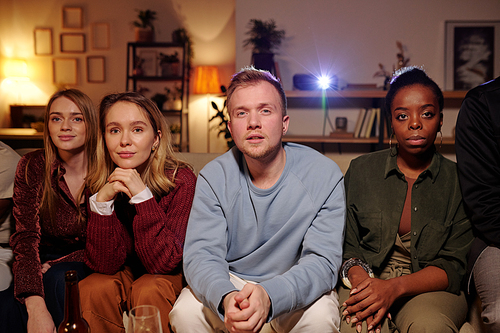 Group of young intercultural friends relaxing on couch in living-room and watching interesting movie together while enjoying home party