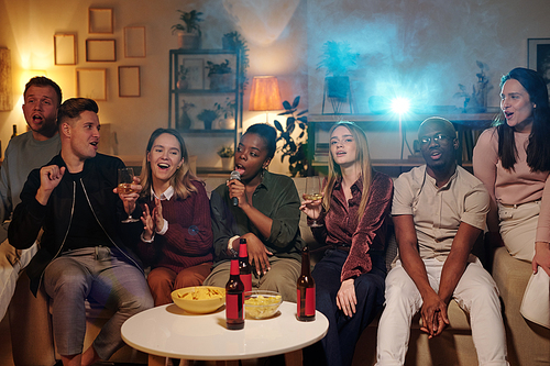 Group of young intercultural friends sitting on long couch by small table with snacks and drinks and singing karaoke together in living-room