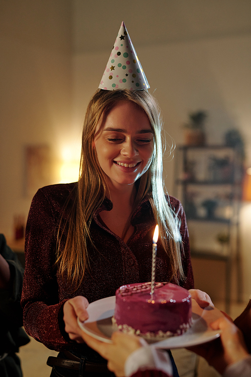 Happy gorgeous blond girl looking at delicious glazed birthday cake with burning candle on plate held by young female during home party