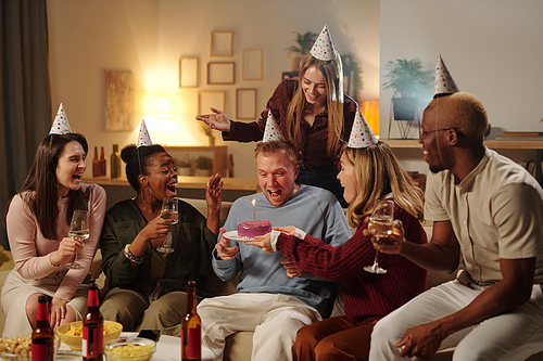 Cheerful young intercultural friends congratulating excited guy on his birthday looking at burning candle on glazed delicious cake