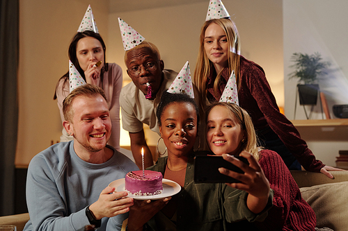 Cheerful young intercultural friends congratulating smiling guy on his birthday while looking at smartphone camera and making selfie