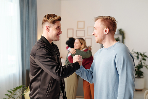 Two young happy men in casualwear standing in front of one another during arm wrestling at home party against girls in embrace