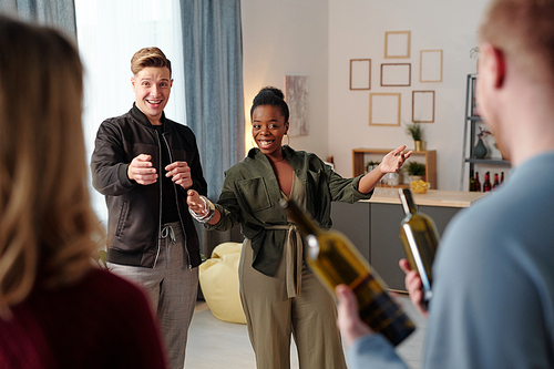 Joyful young intercultural couple meeting their friends with bottles of wine while guy talking to blond woman and inviting guests to come in