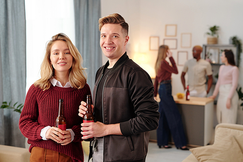 Happy young affectionate couple with bottles of beer looking at you with toothy smiles while enjoying home party against chatting friends