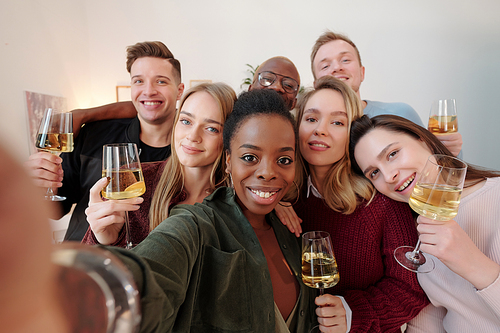 Large group of happy young intercultural friends in smart casualwear making selfie while cheering up with flutes of champagne at party