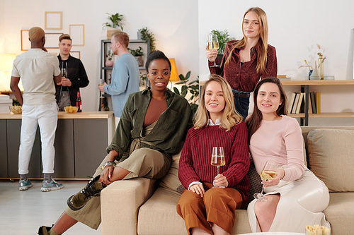 Happy young intercultural women with flutes of champagne looking at you with toothy smiles while enjoying home party against their boyfriends