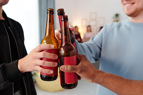 Hands of young intercultural men in casualwear clinking with bottles of beer while having fun at home party and toasting for friendship