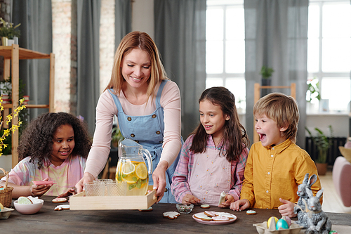 Young woman putting tray with lemonade on the table and treating kids with drink and cookies