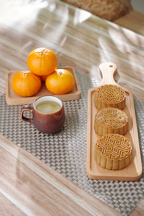 Wooden board with delicious mooncakes, cup of herbal tea and ripe tangerines on kitchen table