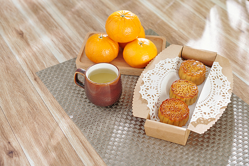 Box of glazed moon cakes, cup of herbal tea and plate with ripe tangerines on kitchen table