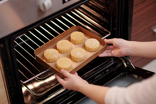 Hands of woman putting tray of decorated mooncakes in preheated oven