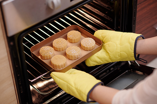 Woman in baking gloves putting tray with mooncakes in preheated oven when cooking delicious dessert at home
